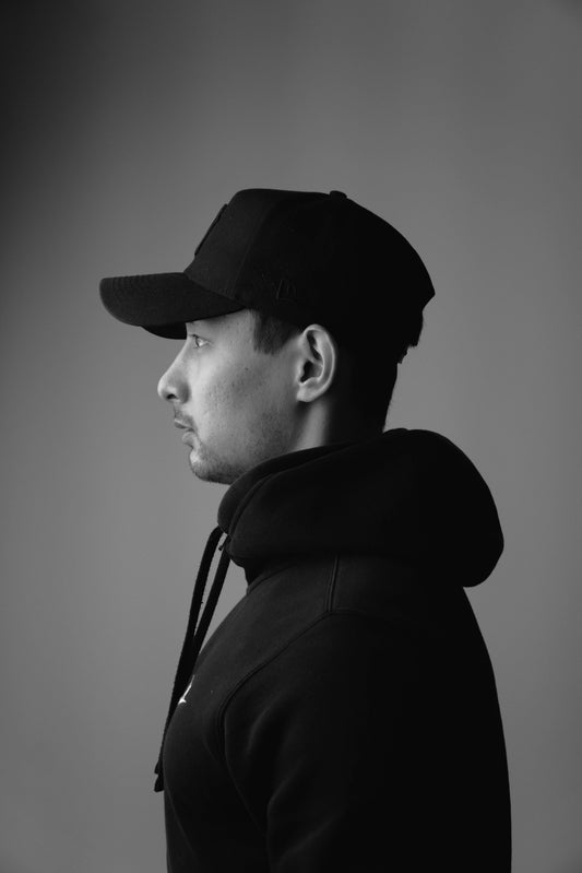 A man in profile wearing a black baseball cap and a black hoodie, photographed in monochrome against a dark gray studio background.